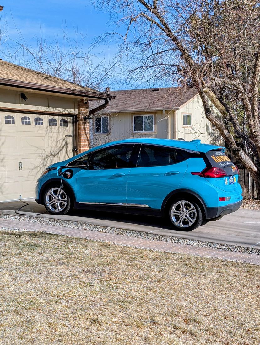 The turquoise Chevy Bolt parked in a driveway, plugged in and charging.