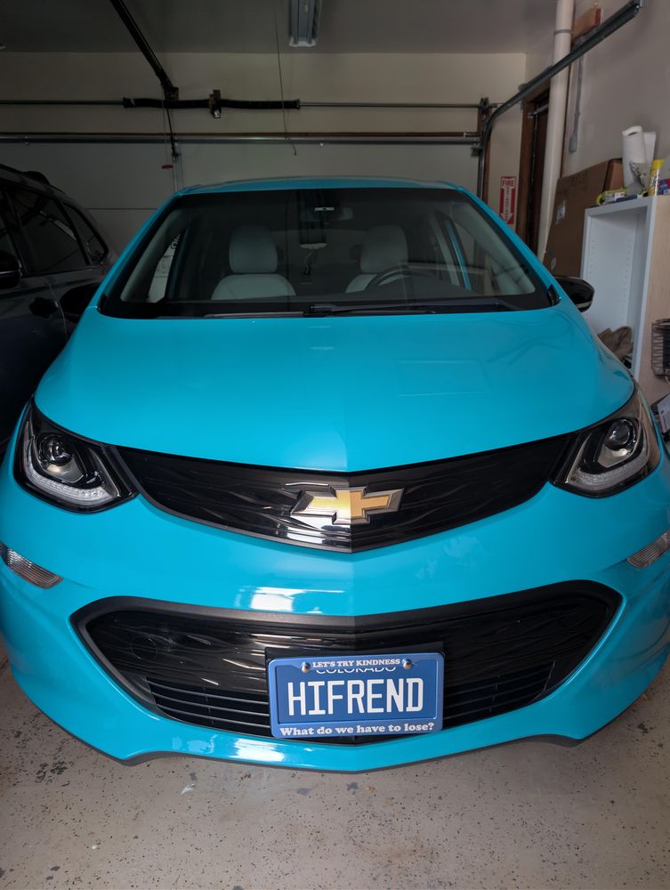 Front of the turquoise Chevy Bolt showing the HIFREND front license plate in a frame that reads 'Let's try kindness' above and 'What do we have to lose?' below.
