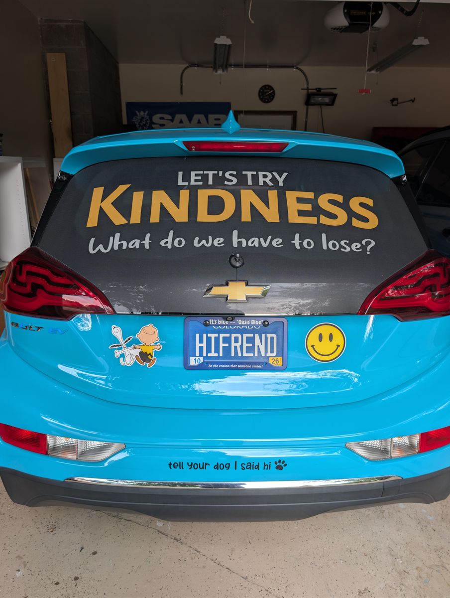 Rear window of a turquoise Chevy Bolt reading 'Let's try kindness. What do we have to lose?' above a Colorado license plate that says HIFREND, flanked by a smiley face sticker and a Charlie Brown sticker.